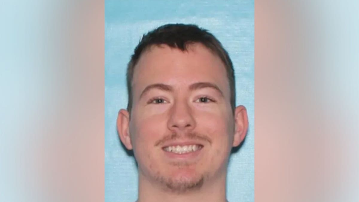 A booking-style ID photograph of a young man with short hair and a goatee against a light blue background.