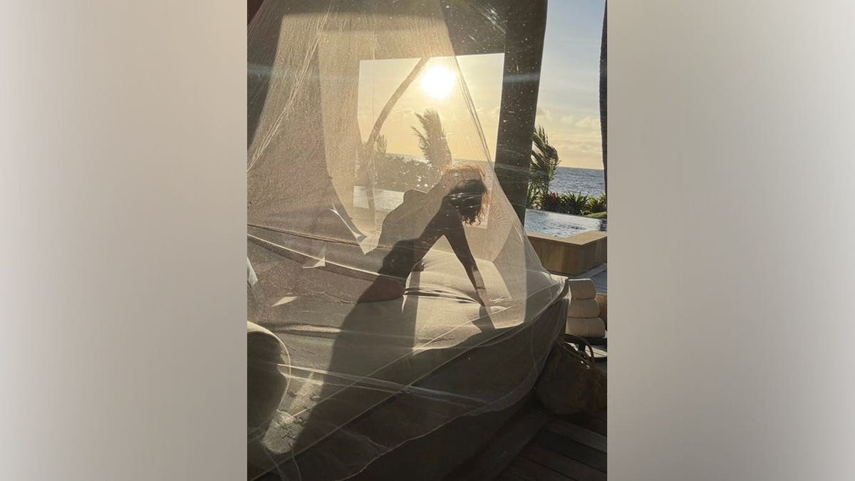 Halle Berry poses in silhouette beneath sheer draped fabric on an outdoor daybed overlooking an infinity pool, palm trees and the ocean during a sunset in Fiji.