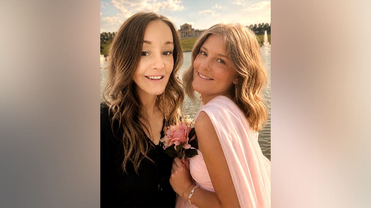 A woman and a teenage girl smile together outdoors near a fountain, the girl holding a pink rose.