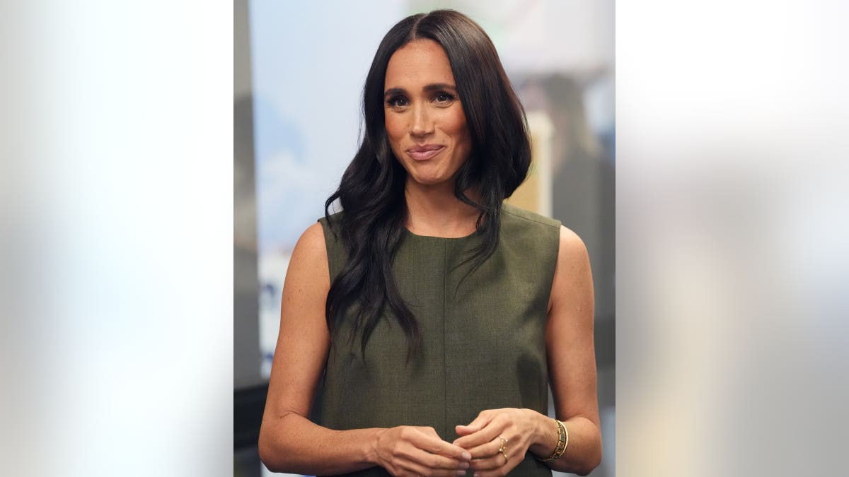 Meghan Markle wears an olive-green sleeveless dress and smiles during a public appearance.