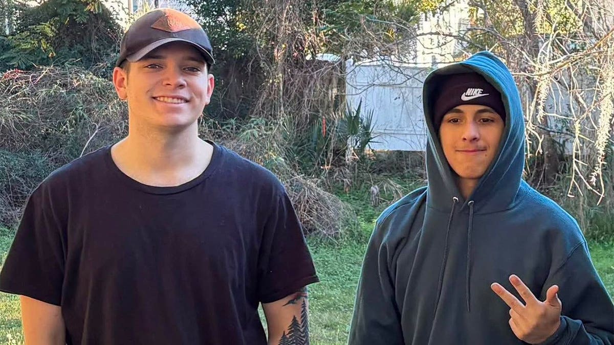 Two young men stand smiling side by side in a backyard, one in a dark T-shirt and cap, the other in a hooded sweatshirt flashing a peace sign.