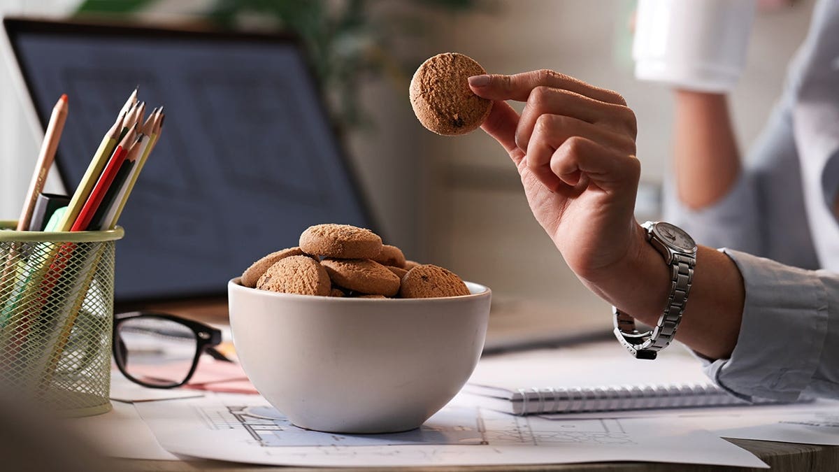 Businesswoman takes a cookie from a bowl while working in an office.