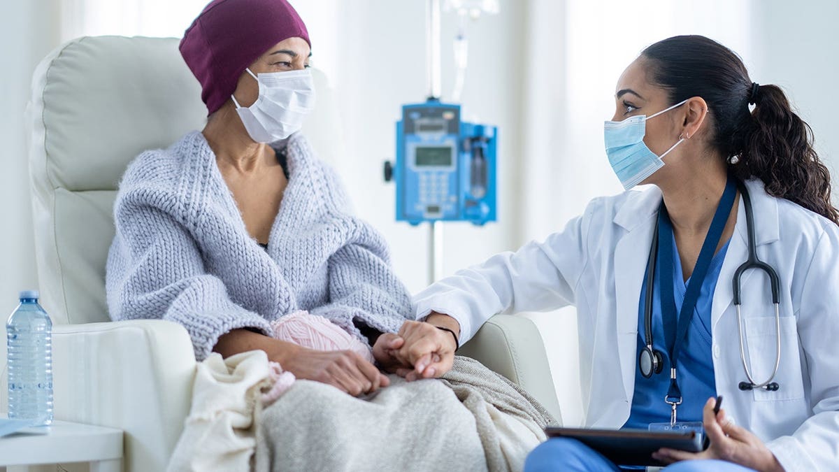 Senior woman battling cancer sits in high-back chair talking with her oncologist during chemotherapy treatment.