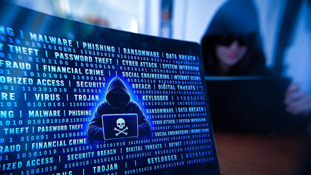 Hooded person uses a digital tablet beside a laptop displaying a pirate symbol in a dark environment.