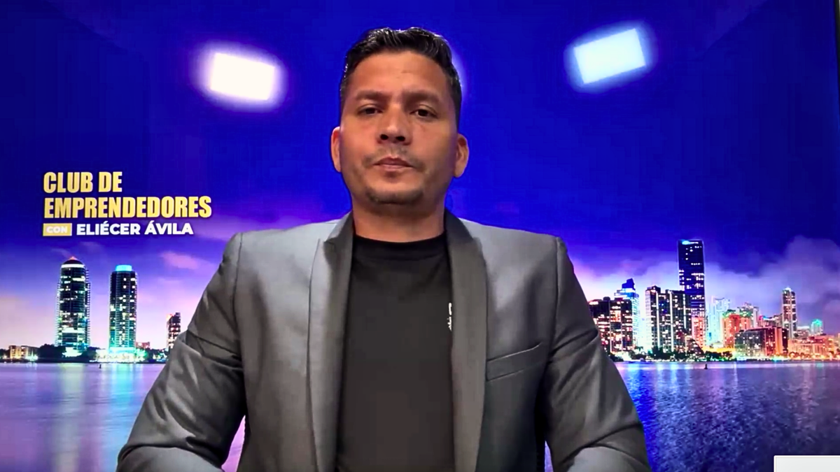 Cuban man sits in front of blue background while looking at computer camera during Zoom interview