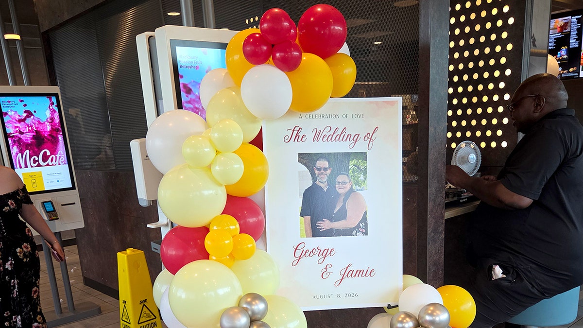 A sign reads "A celebration of love, the wedding of George & Jamie, August 8, 2026," inside a McDonald's restaurant.