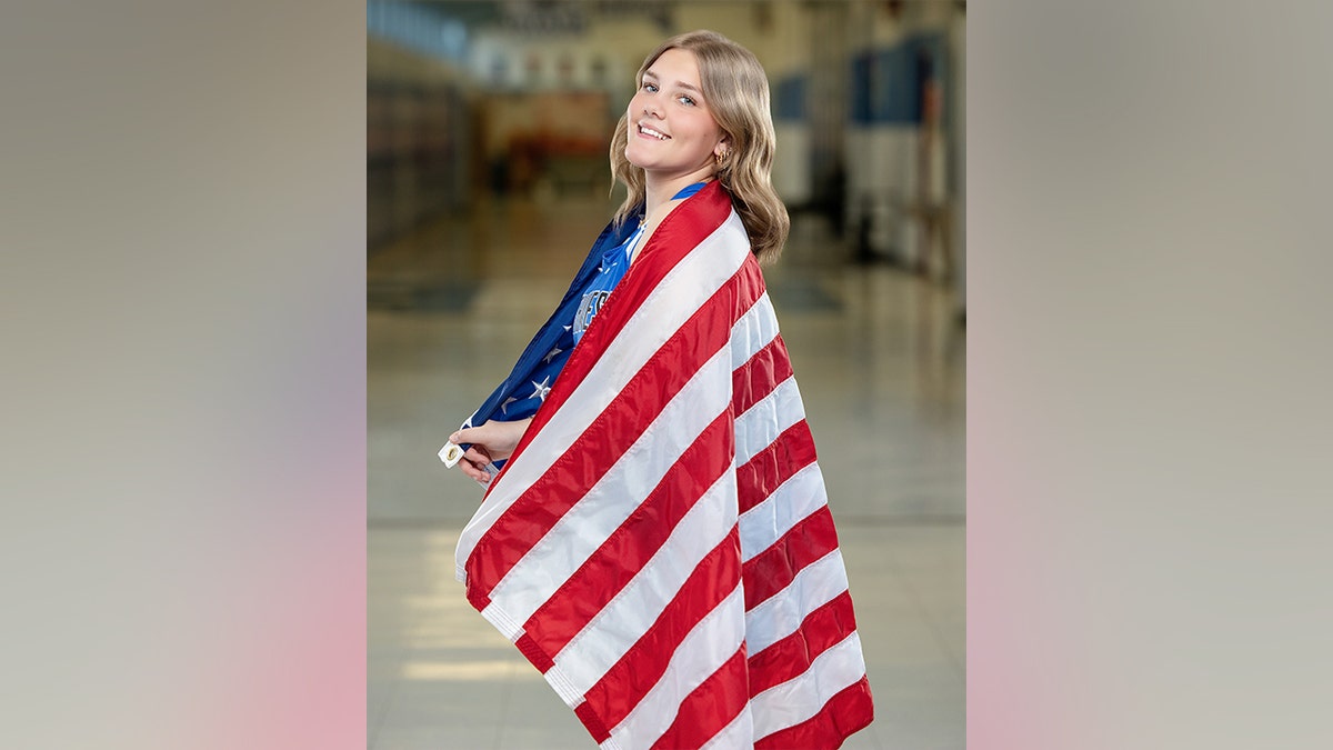 Una adolescente con un uniforme azul de lacrosse del Northwest n.º 30 sostiene una bandera estadounidense extendida a sus espaldas en el pasillo de un colegio.