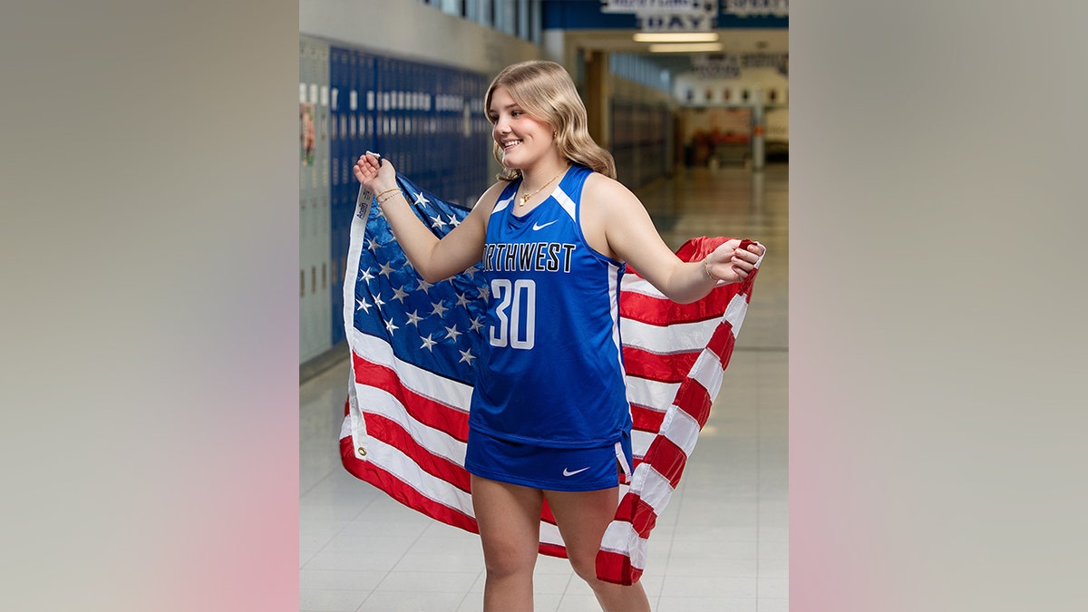 Una adolescente sonríe por encima del hombro, envuelta en una bandera estadounidense que cubre su camiseta deportiva azul, en el pasillo de un colegio.