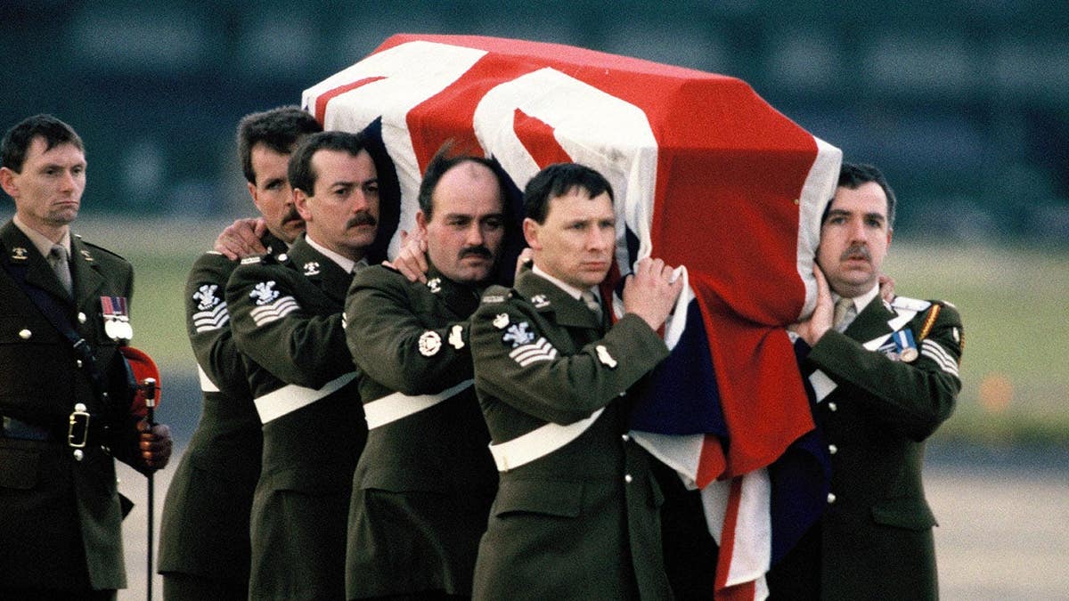 Major Hugh Lindsay's draped coffin is carried by soldiers at RAF Northolt as Prince Charles, Princess Diana and Sarah Ferguson observe.