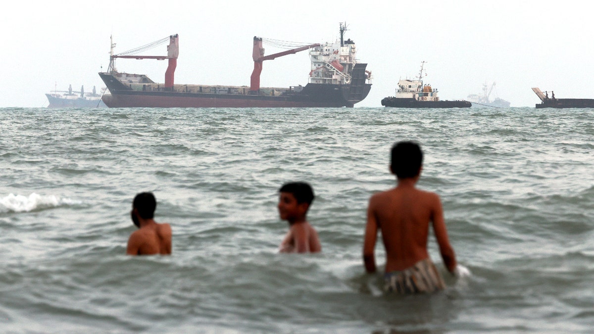 Children swim as ships are seen anchored in the Strait of Hormuz on August 10, 2026, off the coast of Bandar Abbas, Iran.