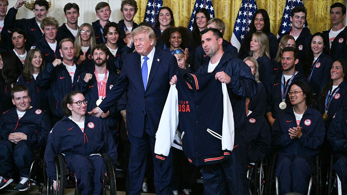 Laurie Stephens smiles as Frank Del Duca presents President Trump with shoes and a Team USA jacket.