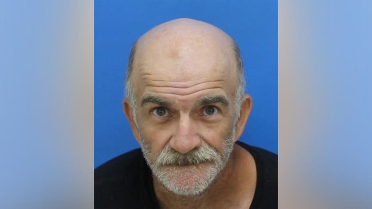 A bald man with a gray mustache and beard wearing a black shirt looks at the camera against a blue background.