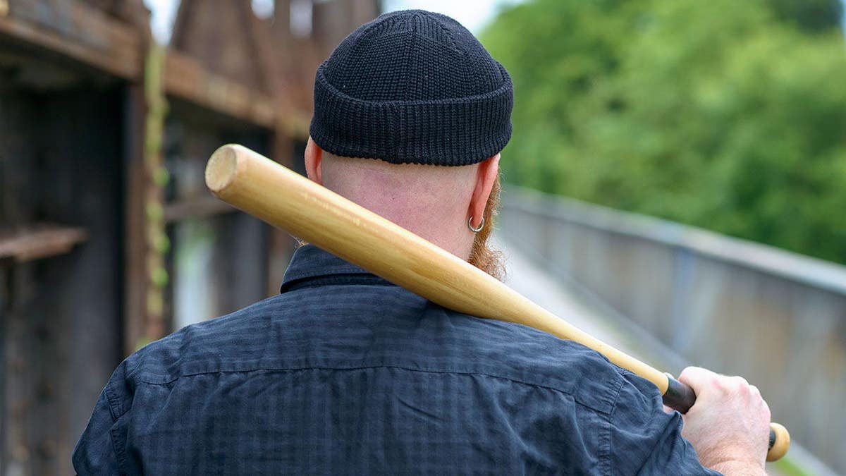 Man wearing gold earring and beanie hat crosses bridge with baseball bat over shoulder, walking away.