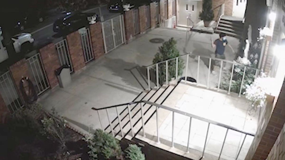 Masked man in dark shirt jumps over fence into church courtyard.
