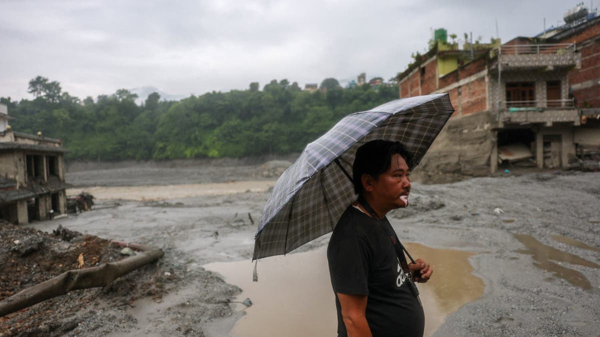 Man holds an umbrella standing before damaged buildings in Trishuli, Nepal.