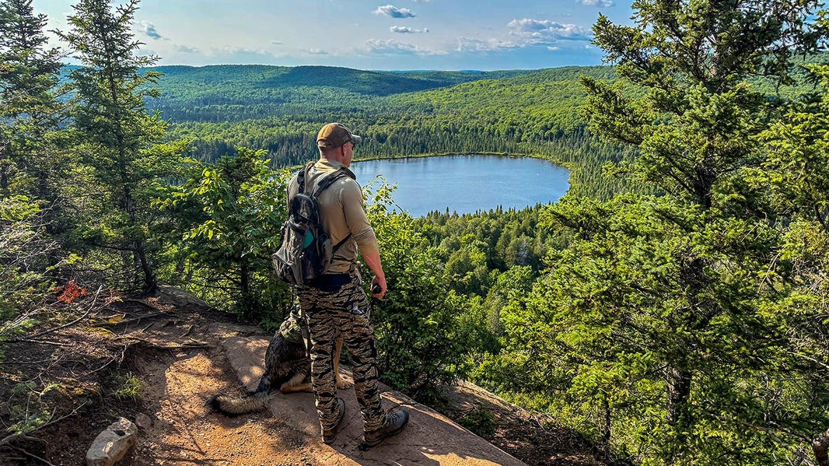 man and dog stand in forest overlooking lake