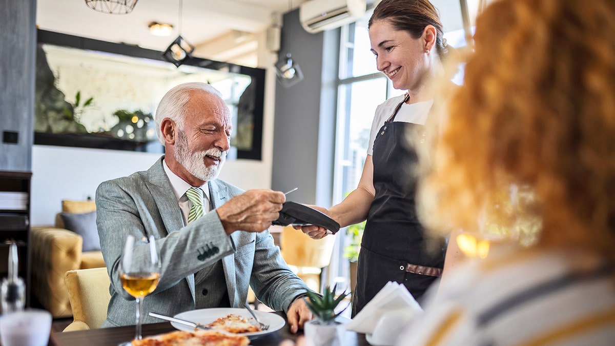 An older man wearing a suit uses a card to pay at a restaurant table.