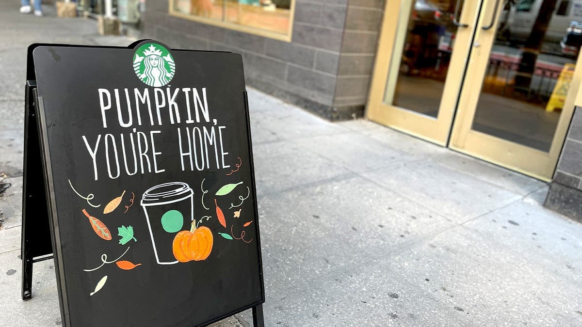 A Starbucks sign advertising pumpkin drinks stands on a sidewalk.