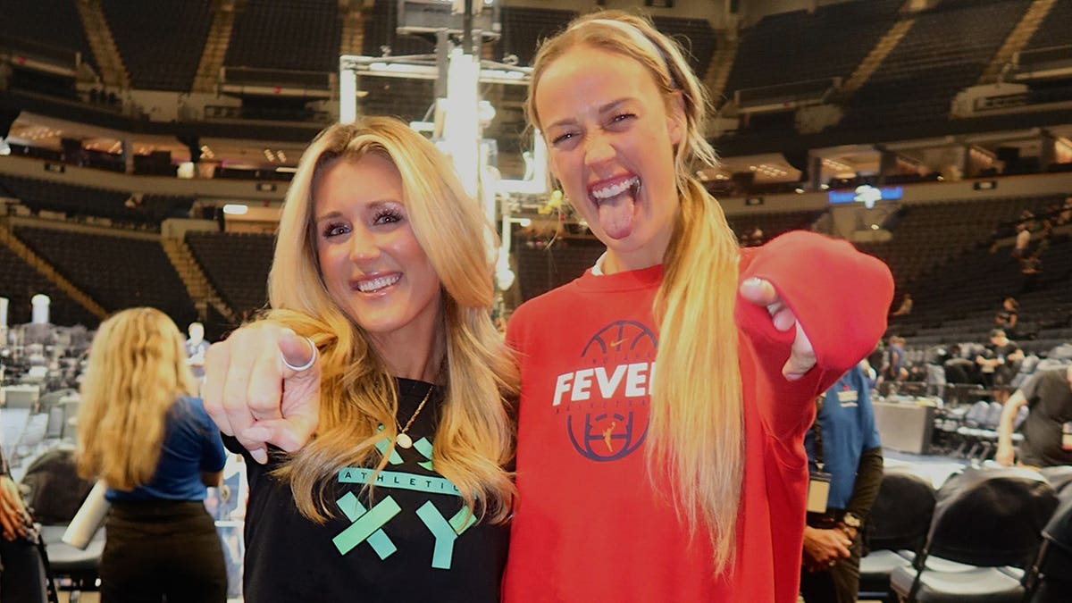 Riley Gaines and Indiana Fever guard Sophie Cunningham posing together on the basketball court, smiling and pointing at the camera inside a packed arena.