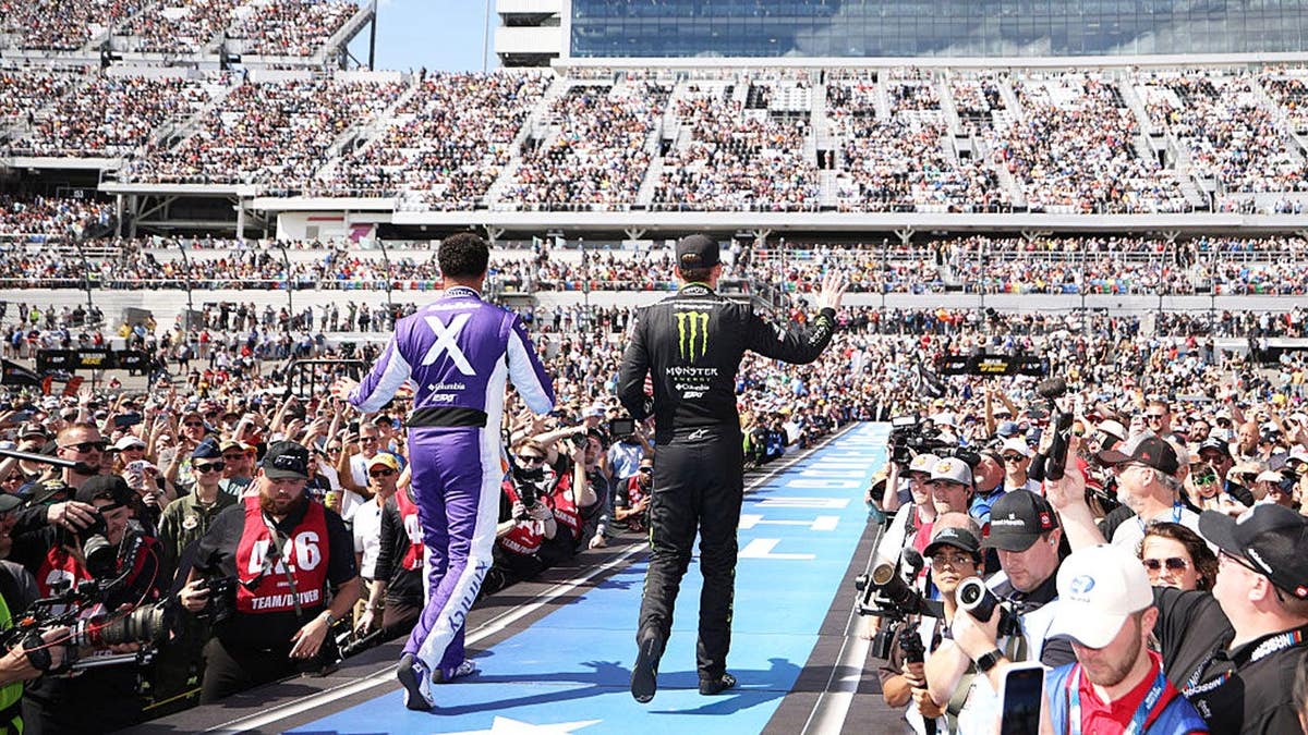 Riley Herbst and Bubba Wallace wave to fans while walking onstage during driver introductions at Daytona International Speedway.