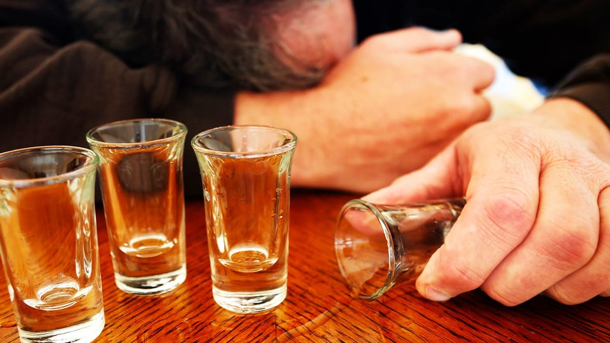 A mature man passed out at a bar from too much drink. Man is laying face down, shot glasses and liquid spilled on the bar.