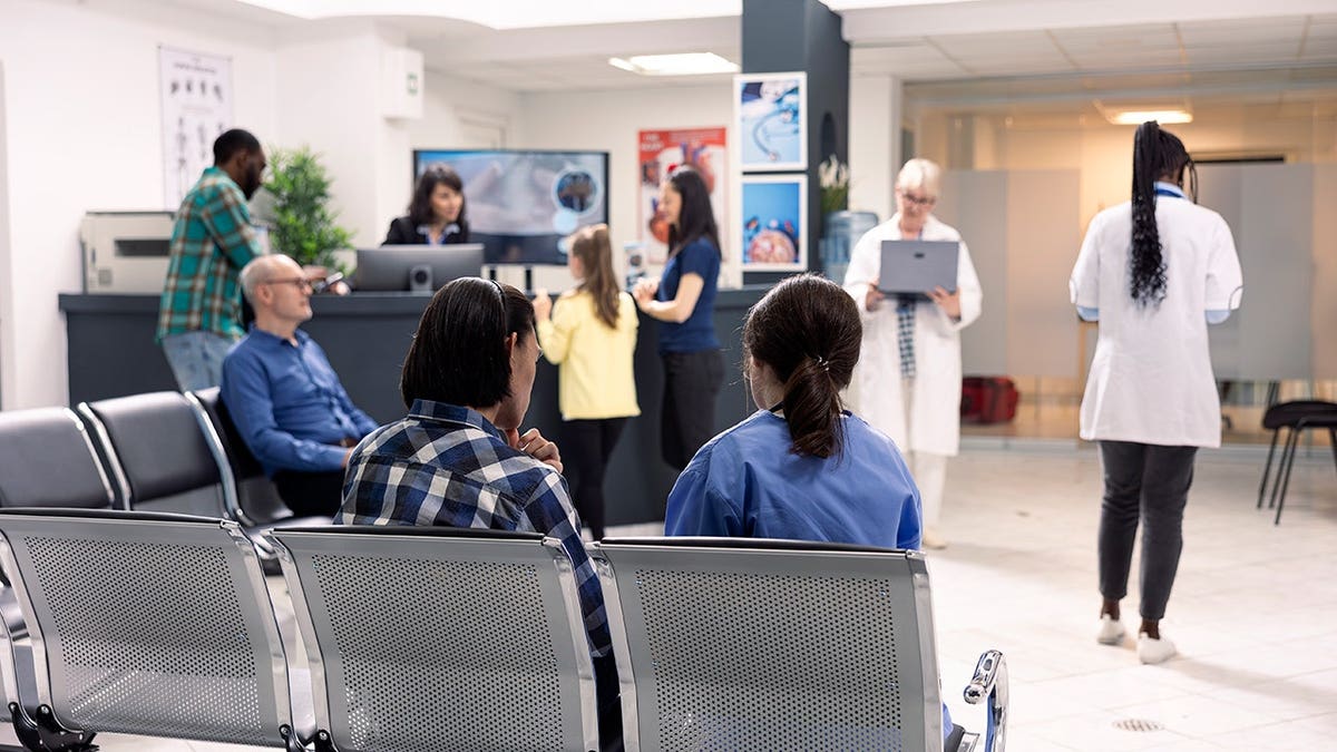 Hospital staff assist patients at front desk in emergency waiting area.