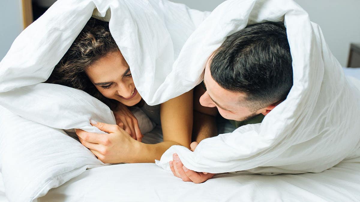Young couple sits together under a blanket on a bed.