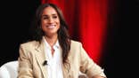Meghan Markle wearing a beige blazer and smiling in front of a red velvet curtain.