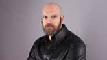 Tyler Mane posing in a leather jacket against a light purple backdrop.