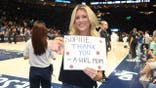 Riley Gaines holding a "Sophie, Thank You – A Girl Mom" sign while standing courtside at an Indiana Fever basketball game and pointing toward the camera.