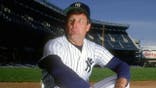 Tommy John in Yankees uniform looks on from the dugout at Yankee Stadium.
