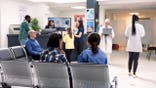Hospital staff assist patients at front desk in emergency waiting area.