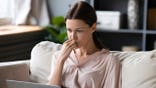 Woman sits on sofa looking puzzled at laptop screen.