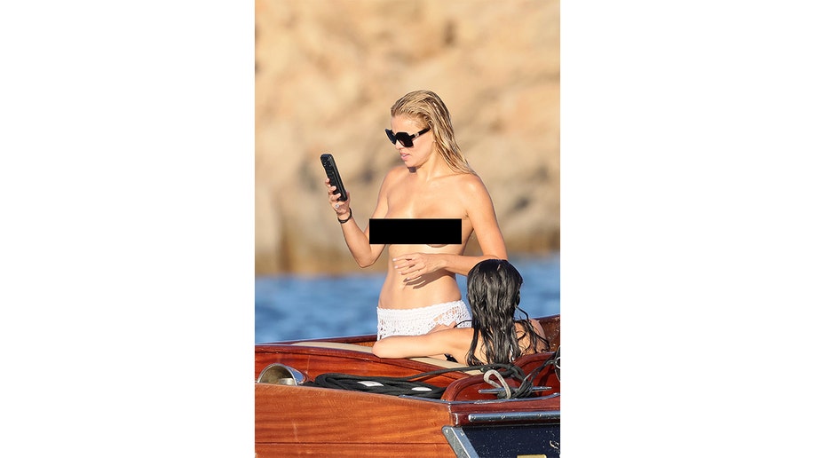 Brooks Nader stands topless in white crochet swim bottoms on a wooden tender, holding a phone.