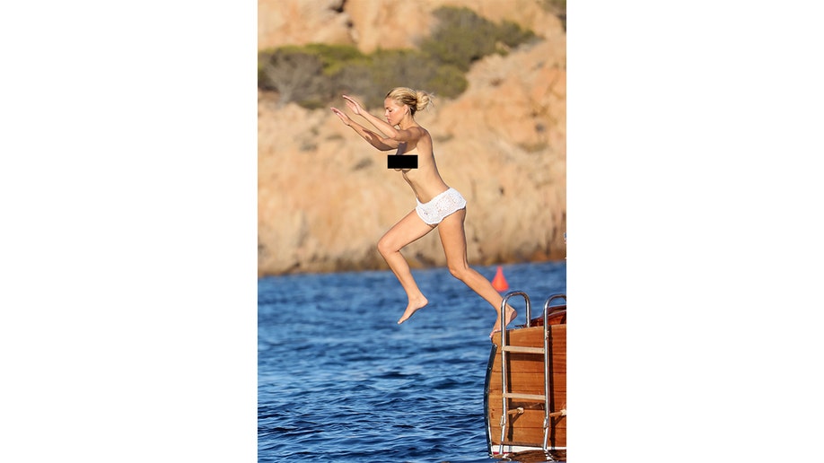 Brooks Nader, topless and wearing white crochet swim bottoms, jumps from a wooden tender into the sea.