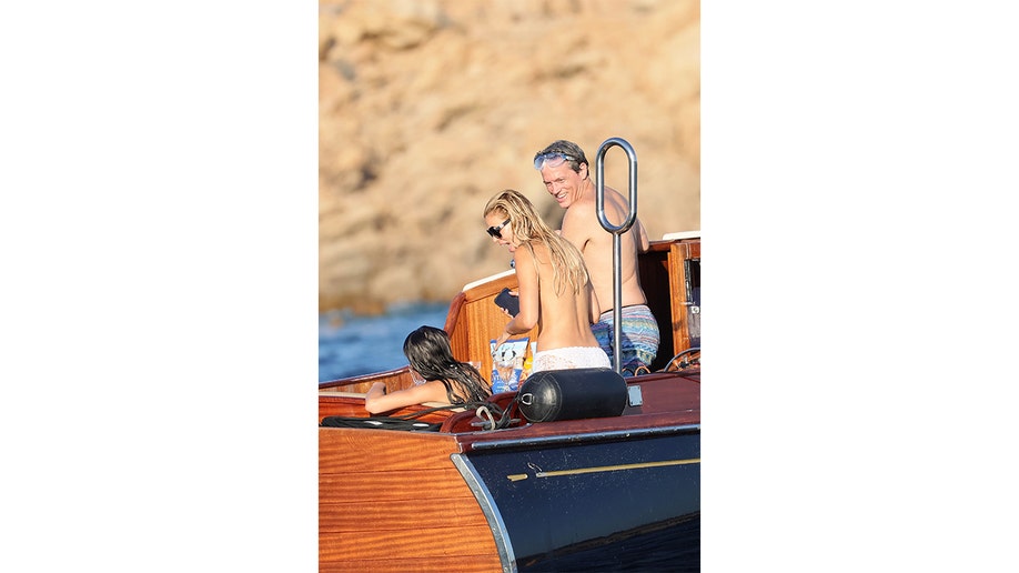 Brooks Nader stands topless, seen from behind, on a wooden tender with two family members nearby.