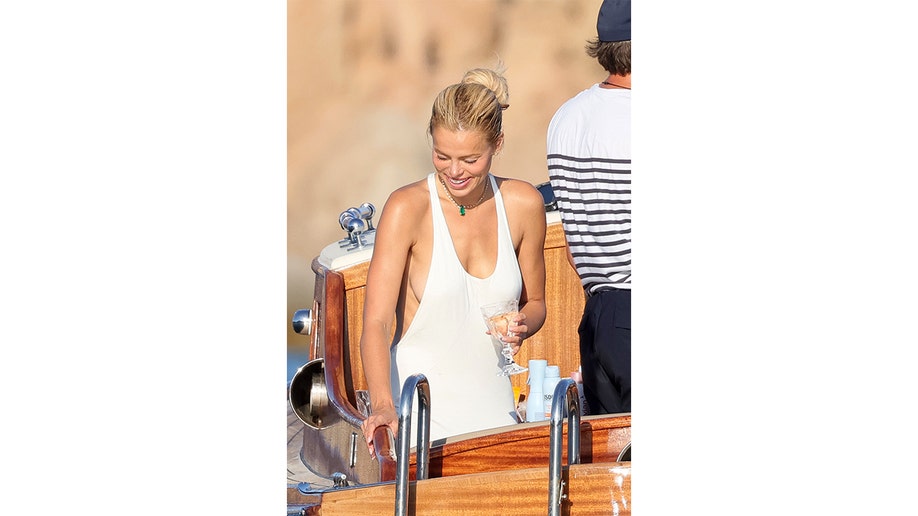 Brooks Nader smiles in a white halter dress while holding a drink aboard a wooden tender.