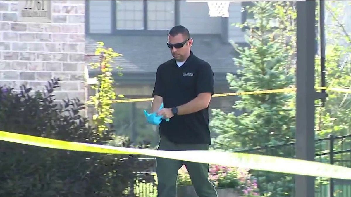 An investigator stands near a doorway behind police tape outside a brick home.