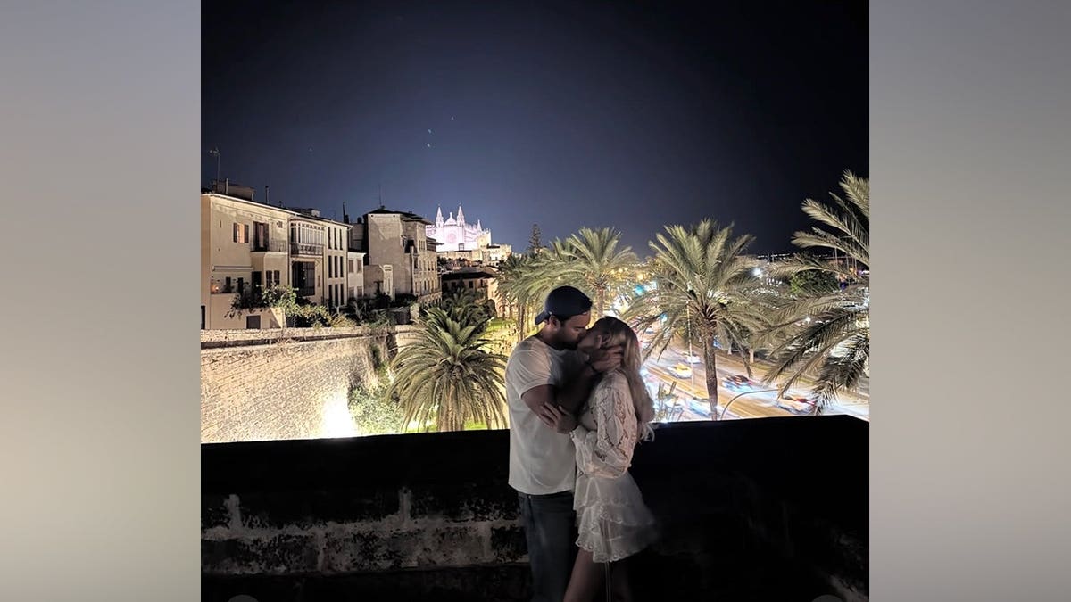 Sydney Sweeney and Scooter Braun kiss while embracing against a palm tree-lined cityscape at night.