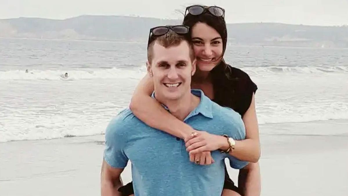 A man and a woman embrace on a beach, both smiling, with ocean waves and a hillside behind them.