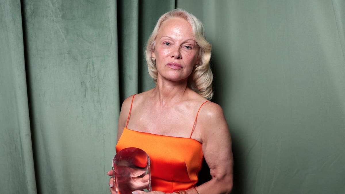 Pamela Anderson wears an orange dress and holds an award while posing indoors.