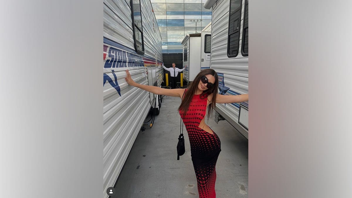 Emily Ratajkowski poses between production trailers while wearing her red-and-black VMAs look and sunglasses.