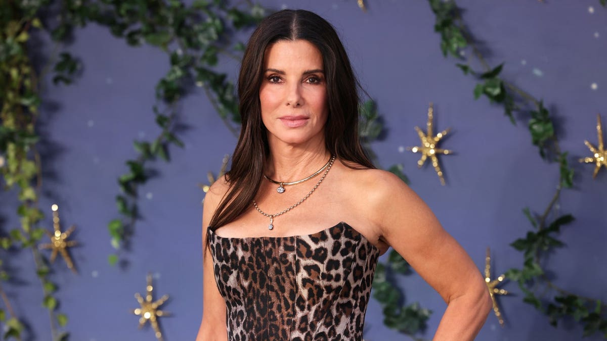 Sandra Bullock poses in a strapless leopard-print dress against a purple floral backdrop.