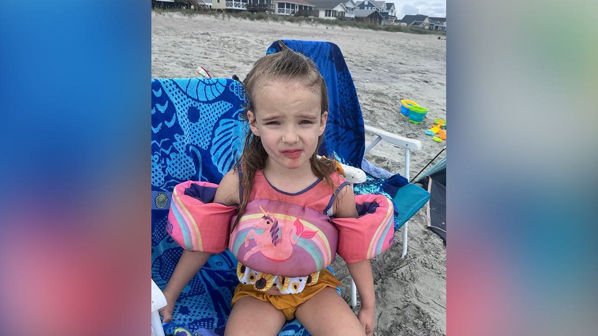 Young girl with brown hair sits on a blue beach chair on a sandy beach while wearing pink flotation devices and yellow shorts.