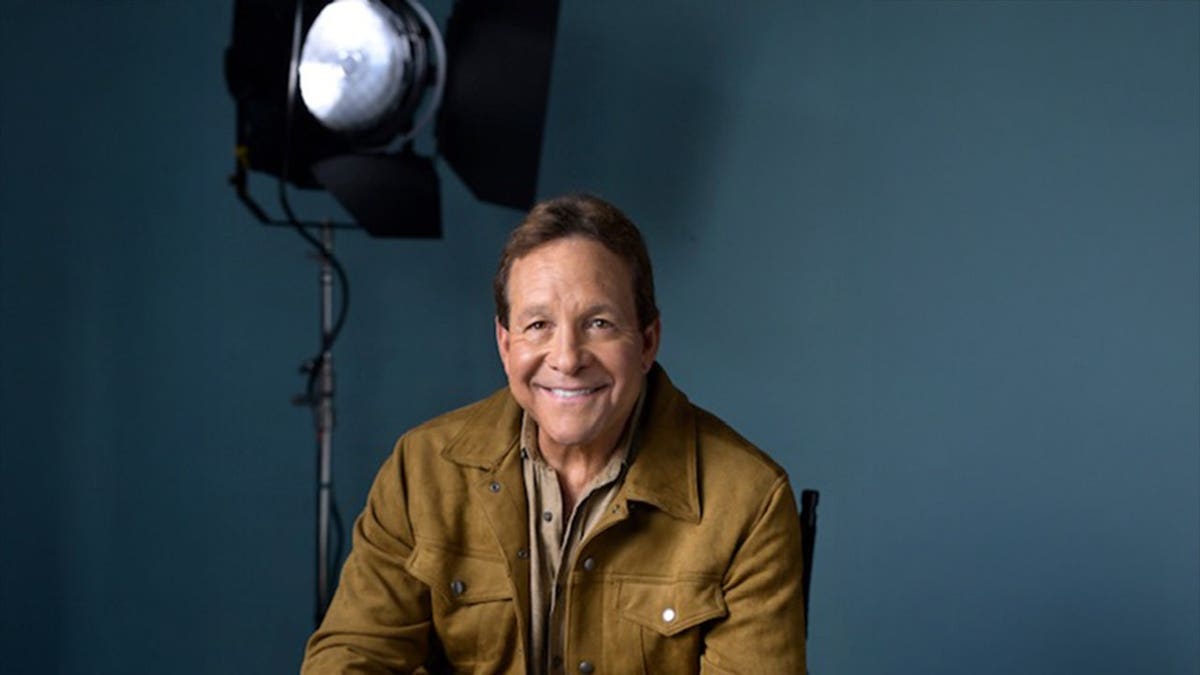 Steve Guttenberg smiling in a brown jacket inside a photo studio.