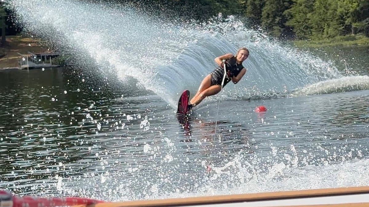 Sydney Sweeney water skis across a lake while wearing a life jacket and holding a tow rope.