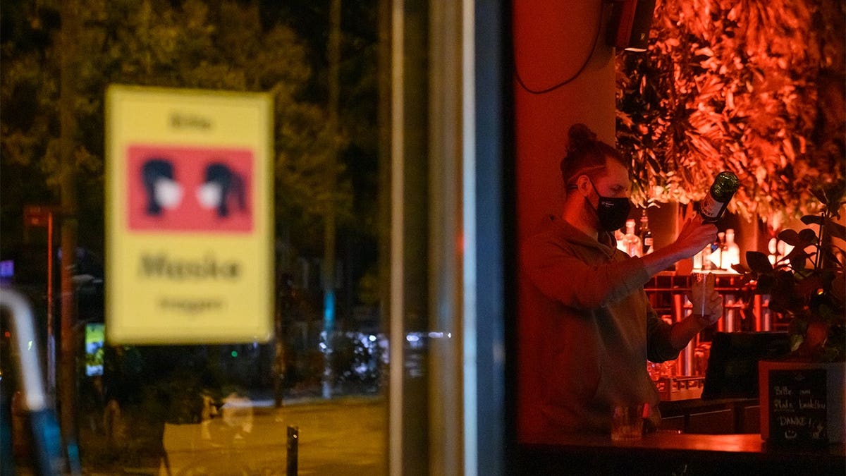 Bartender Ron stands behind a bar counter wearing a mask with a sign reading 'Please wear mask' visible at the entrance.