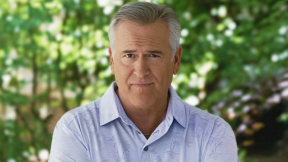 Bruce Campbell poses outdoors with crossed arms while wearing a blue collared shirt.