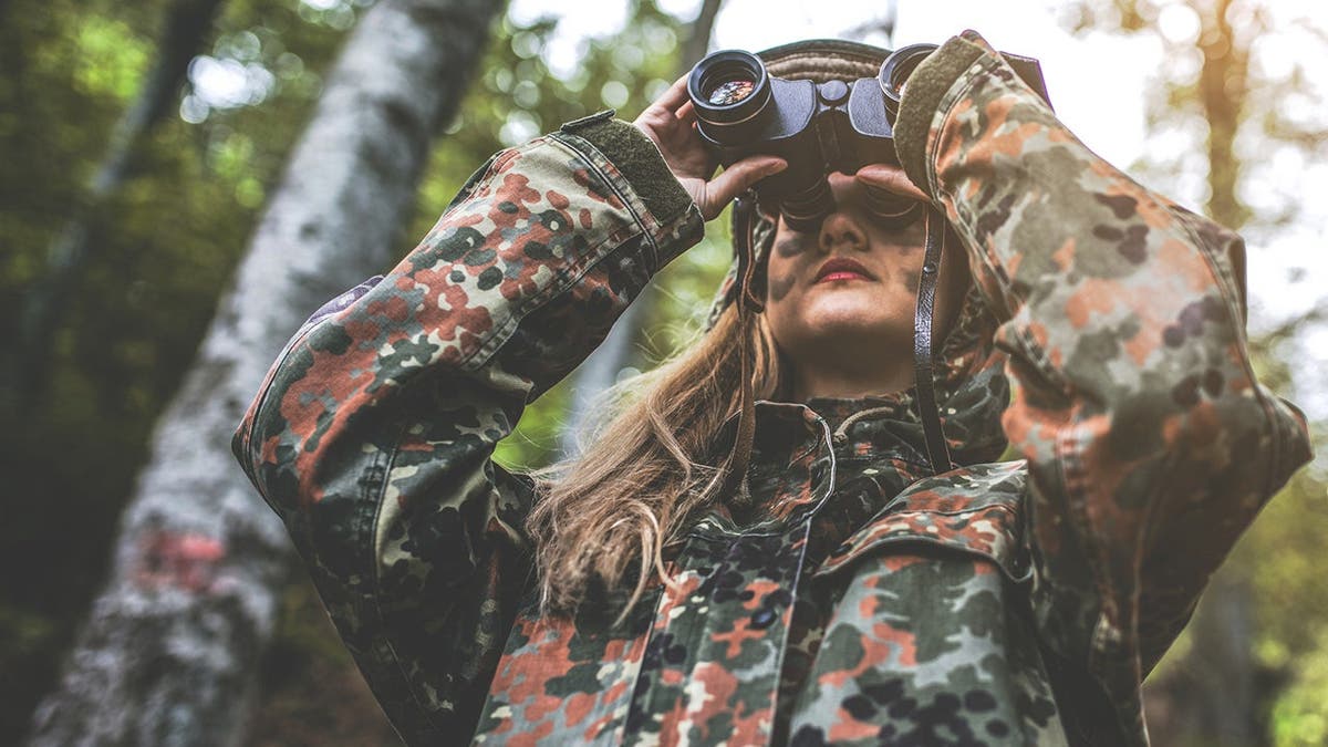 A woman in camouflage stands in a forest.