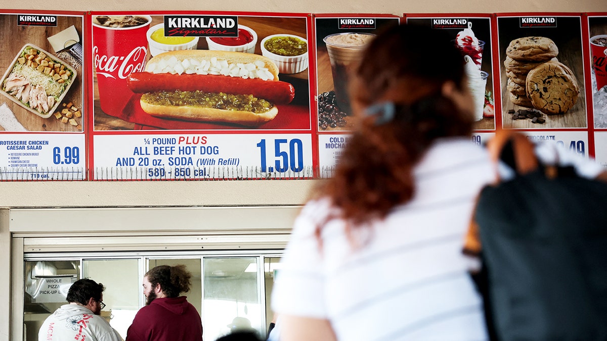 Costco food court customers wait beneath a sign for the Kirkland Signature hot dog with a 20-ounce soda.